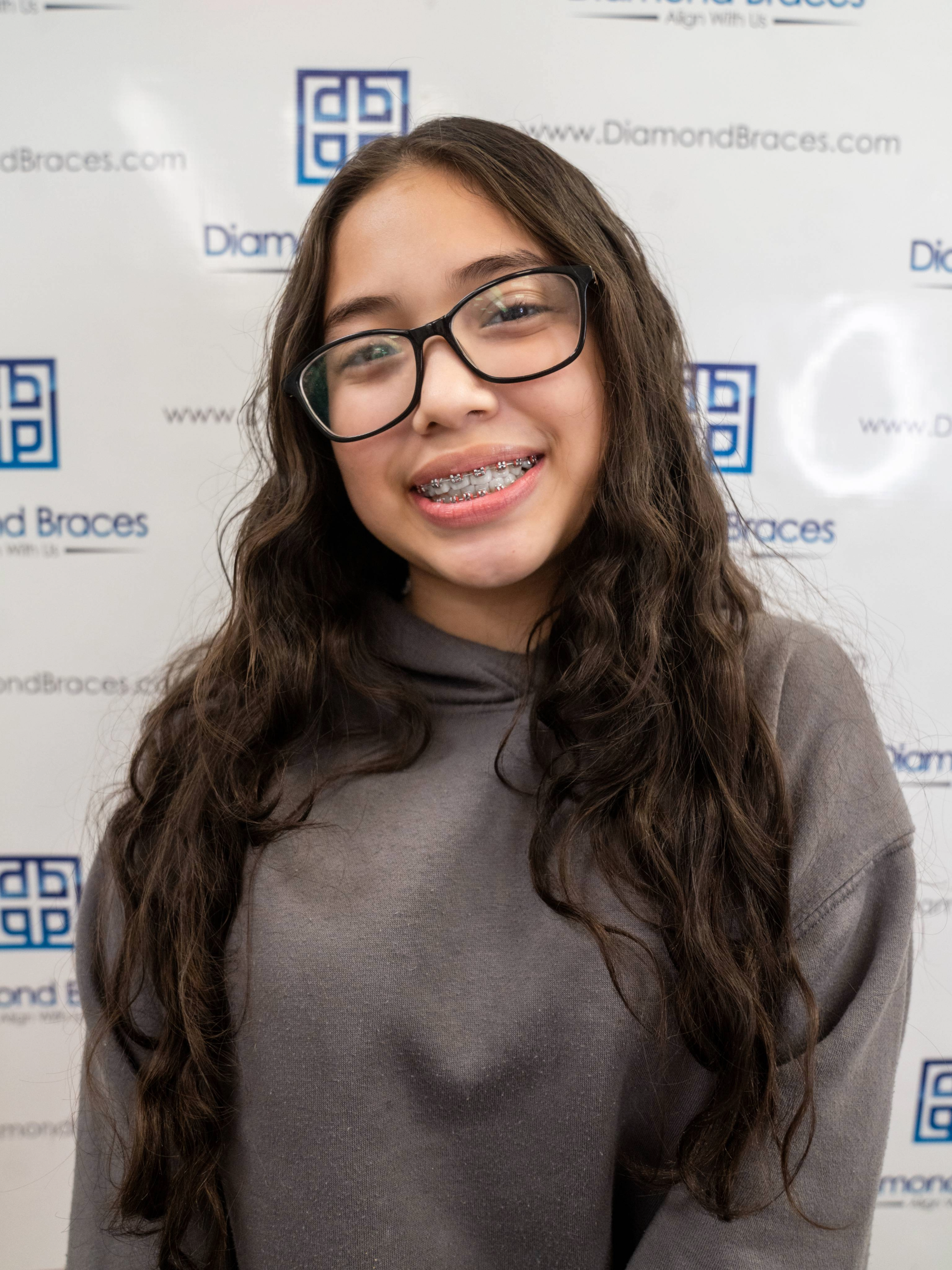 Dental care patient with long hair and glasses standing in front of a Diamond Braces promotional backdrop.