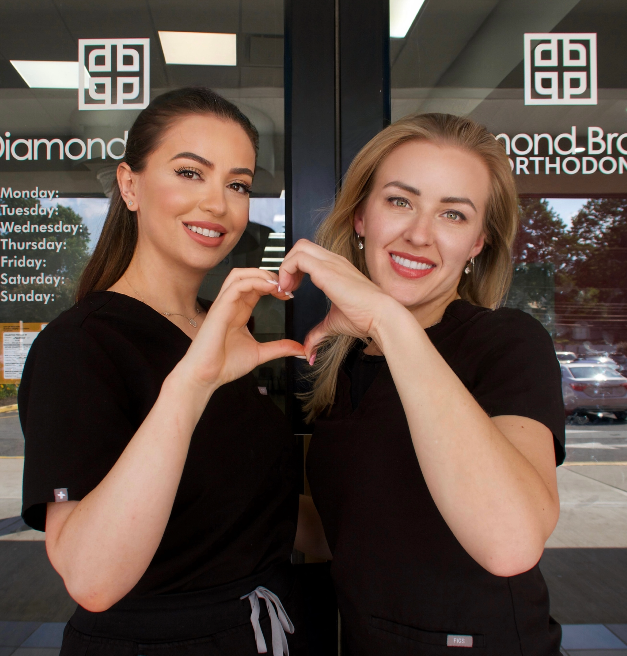 Two Orthodontists in black uniforms making a heart shape with their hands in front of a dental office entrance.
