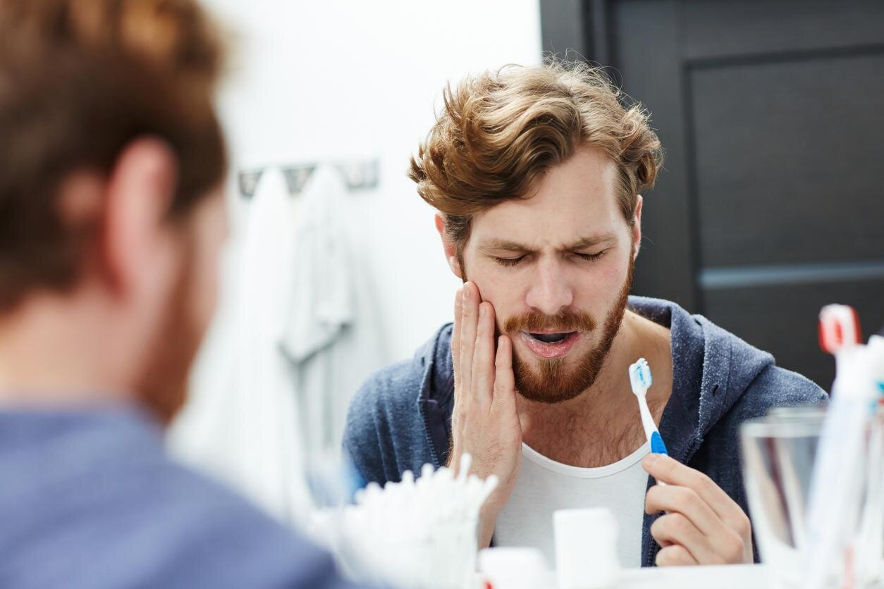 Man in pain in front of the mirror brushing his teeth while he has his hand on the side of his cheek.