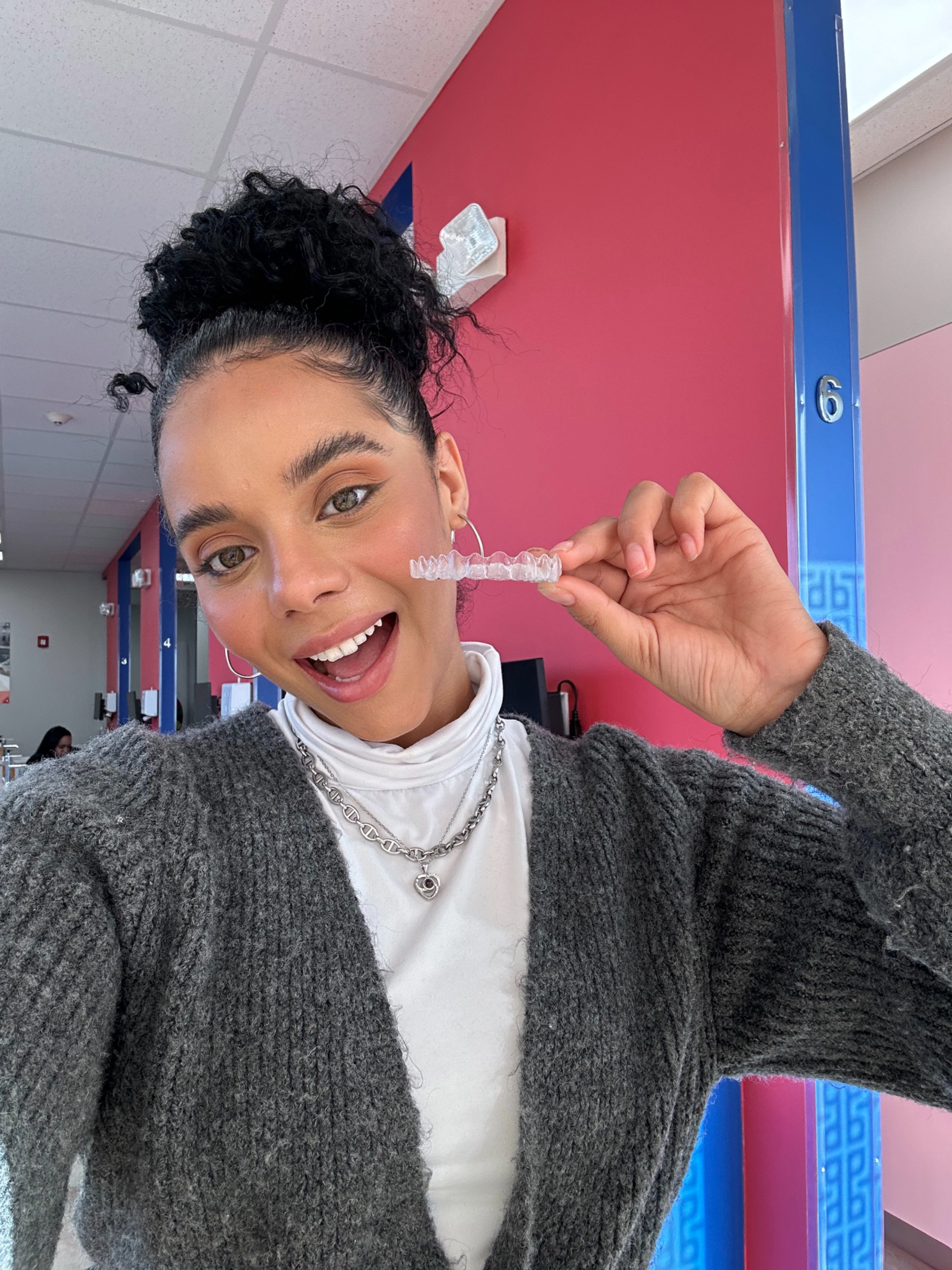 Young dental care patient is smiling big while holding a clear aligner in front of a colorful wall.