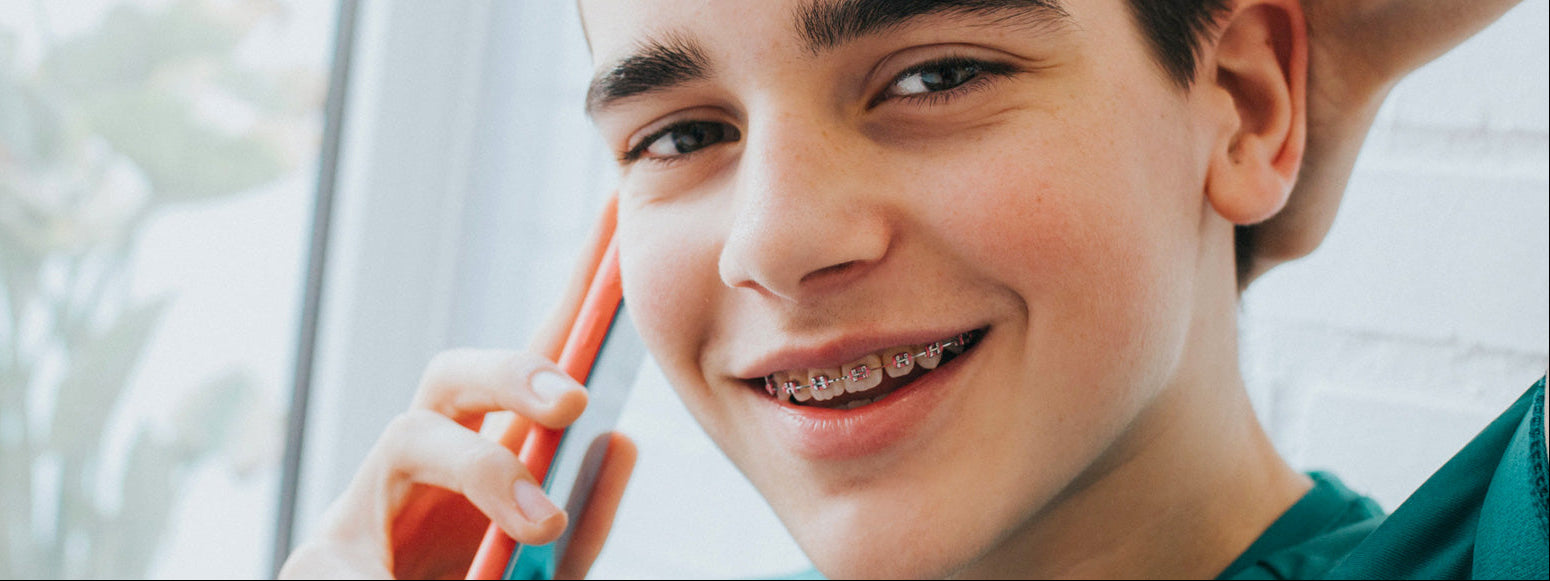 Young boy with metal braces smiling and holding a phone to his ear.