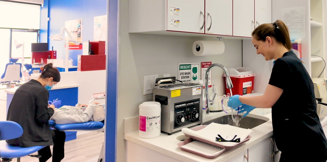 Person washing dishes in a kitchen with another person working at a desk in the background.