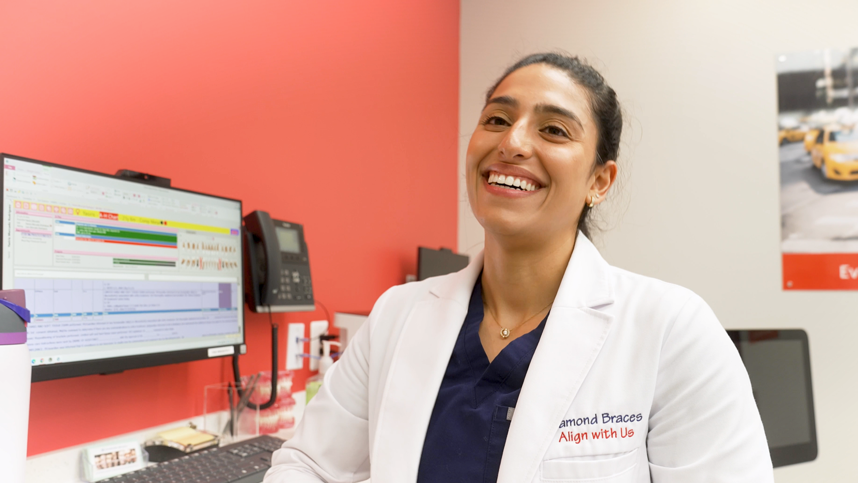 Dentist in a lab coat smiling in a dental office setting with a computer and posters on the wall.