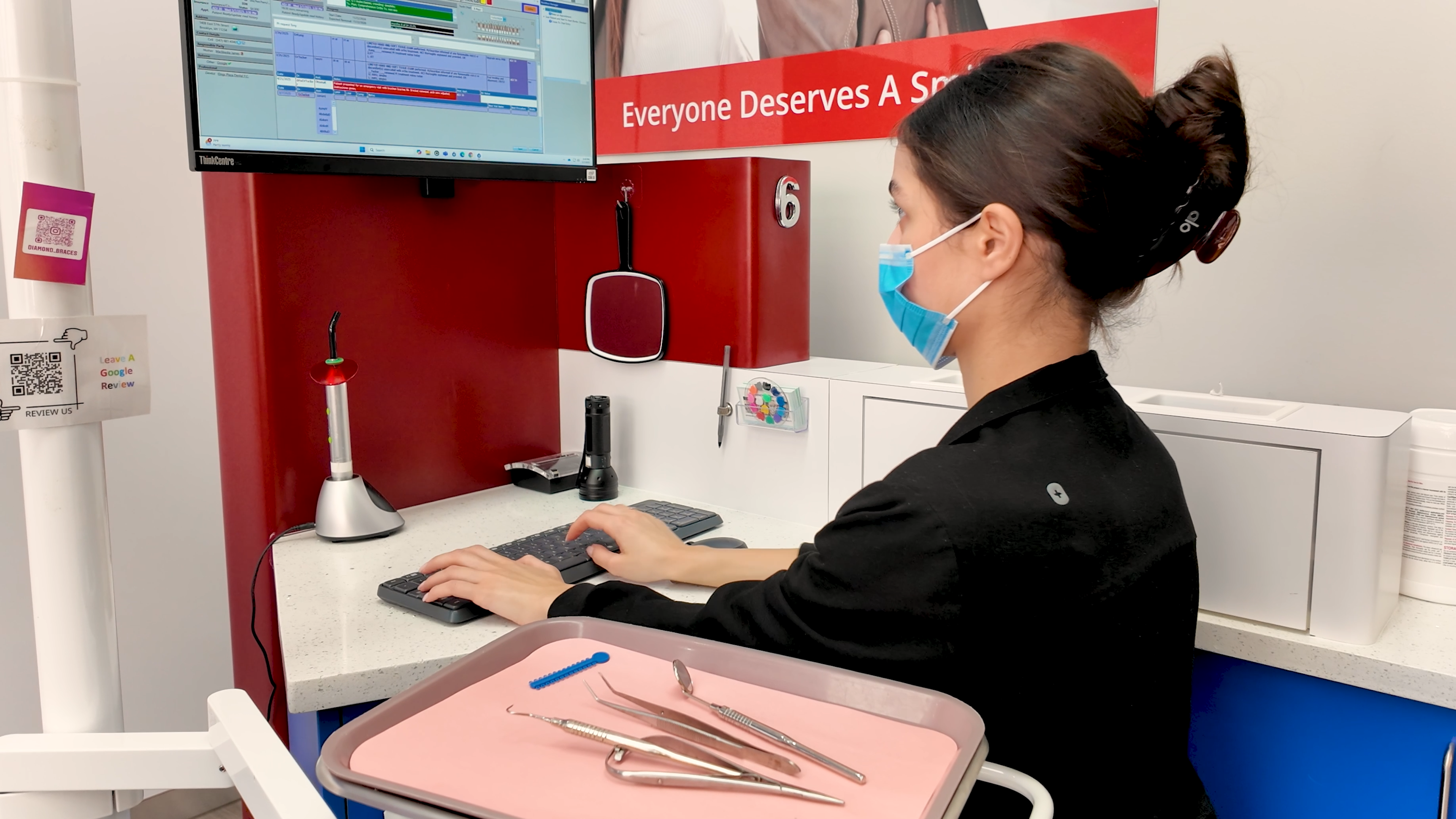 Orthodontic assistant wearing a face mask sitting at a desk with medical tools and computer monitors.
